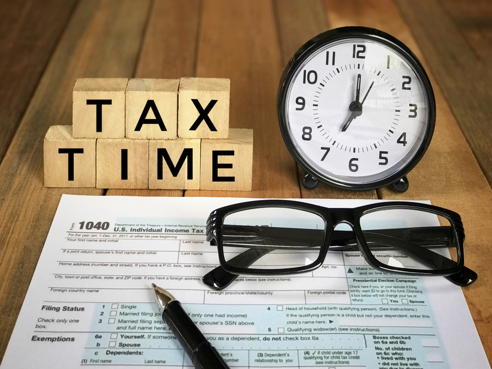 A wooden block spelling 'TAX TIME' beside a clock, eyeglasses, and a filled-out 1040 tax form